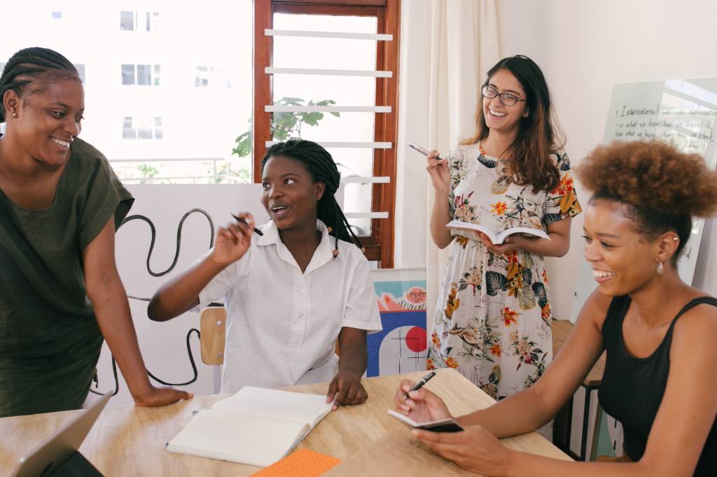 Imagem mostra 4 mulheres, 3 negras e uma branca, sorrindo e conversando em uma mesa de reunião