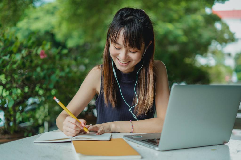 Mulher oriental jovem com fones de ouvidos, estudando atrás de um notebook em um local ao ar livre