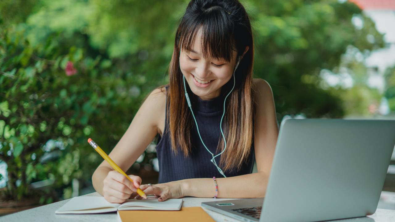 Mulher oriental jovem com fones de ouvidos, estudando atrás de um notebook em um local ao ar livre