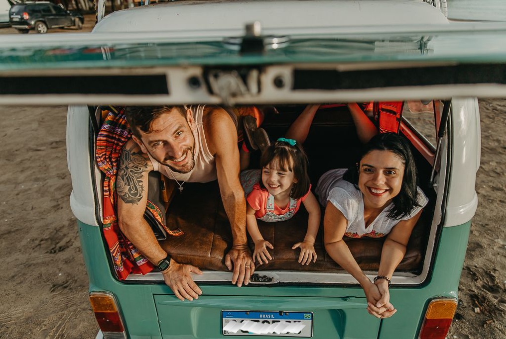 Imagem de uma mulher branca de cabelos escuros, um homem branco de cabelos escuros e uma menina deitados de bruços em uma Kombi verde.