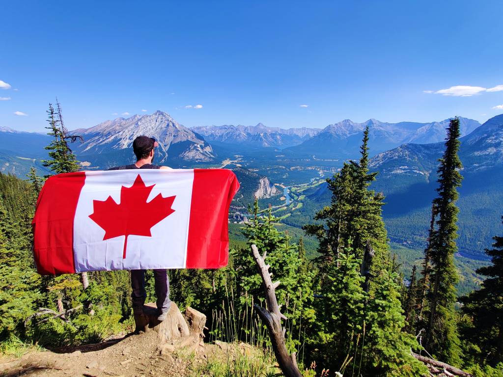 Imagem mostra um homem de costas segurando uma bandeira do Canadá. Ele está no alto de uma montanha.