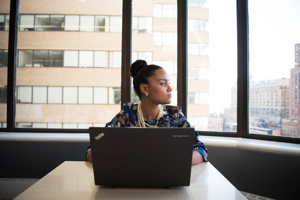 Uma mulher aparece em um escritório, em frente a um computador, olhando para seu lado esquerdo.