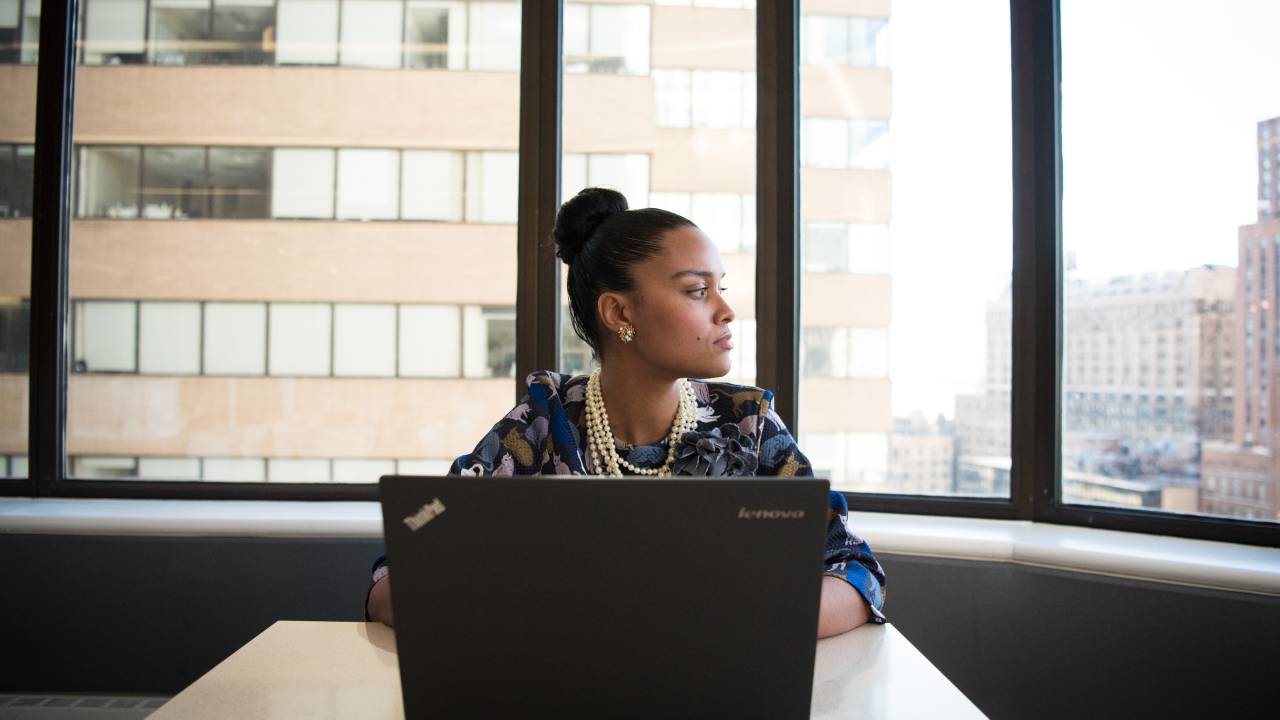 Uma mulher aparece em um escritório, em frente a um computador, olhando para seu lado esquerdo.