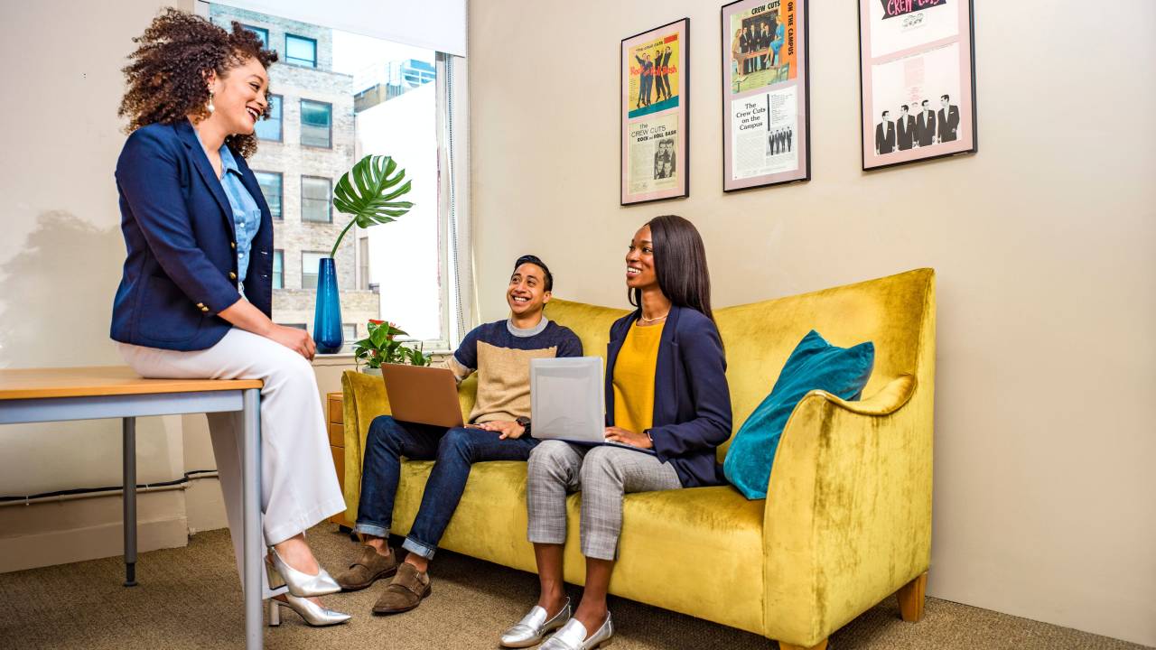 Imagem mostra três pessoas conversando em um escritório: duas mulheres negras e um homem latino