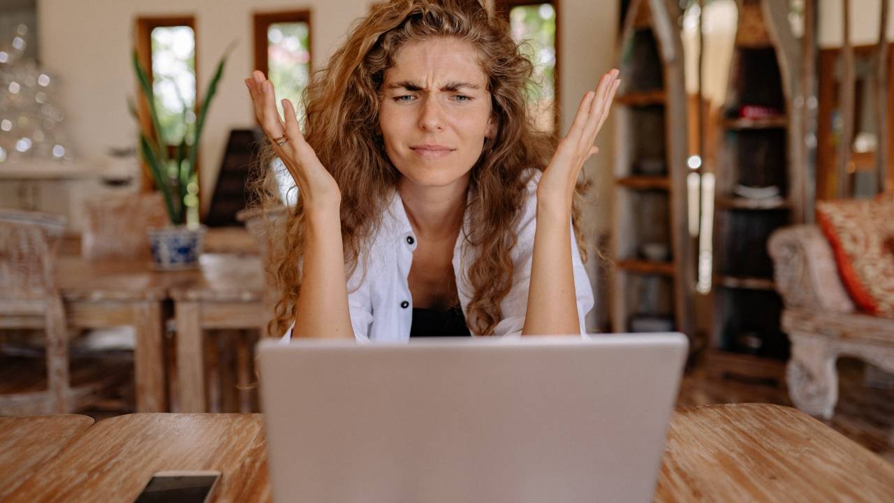 Imagem mostra mulher loira, de cabelos cacheados, fazendo um gesto de descontentamento em frente ao computador.