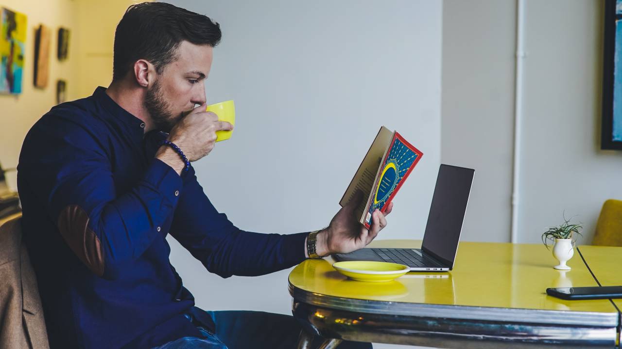 Imagem mostra um homem branco, jovem, sentado numa mesa tomando café e lendo um livro em frente a um notebook
