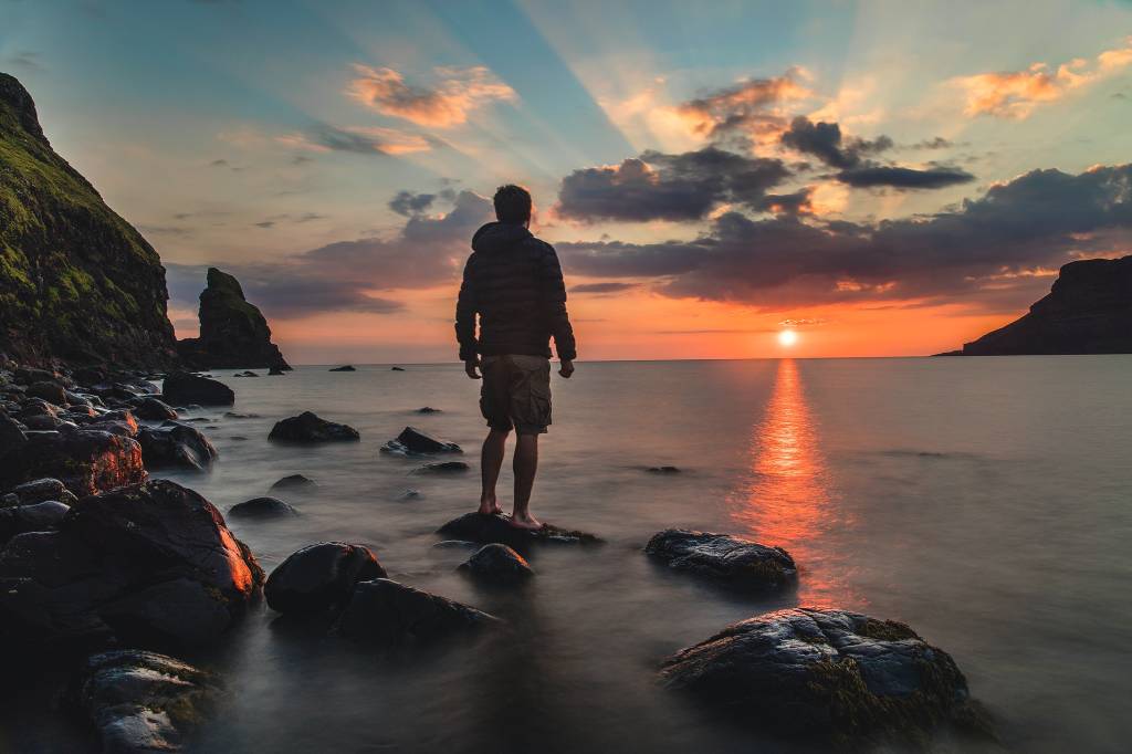 Imagem mostra um home de costas na praia. Ele está parado em cima de uma pedra observando o pôr-do-sol
