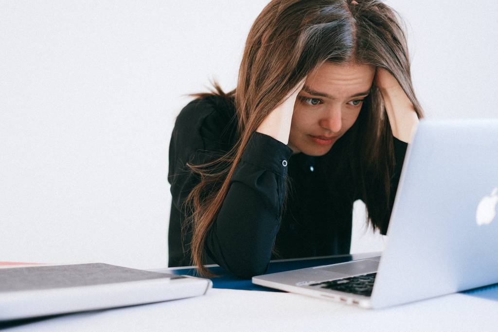 Imagem mostra mulher branca de cabelos castanhos olhando para um computador com as mãos na cabeça e expressão preocupada.