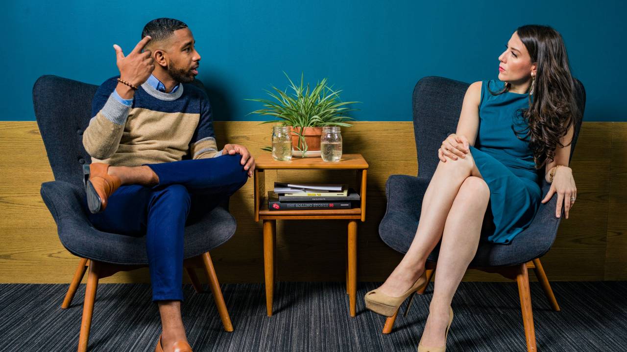 Imagem mostra um homem negro conversando com uma mulher branca. Ambos estão sentados em poltronas