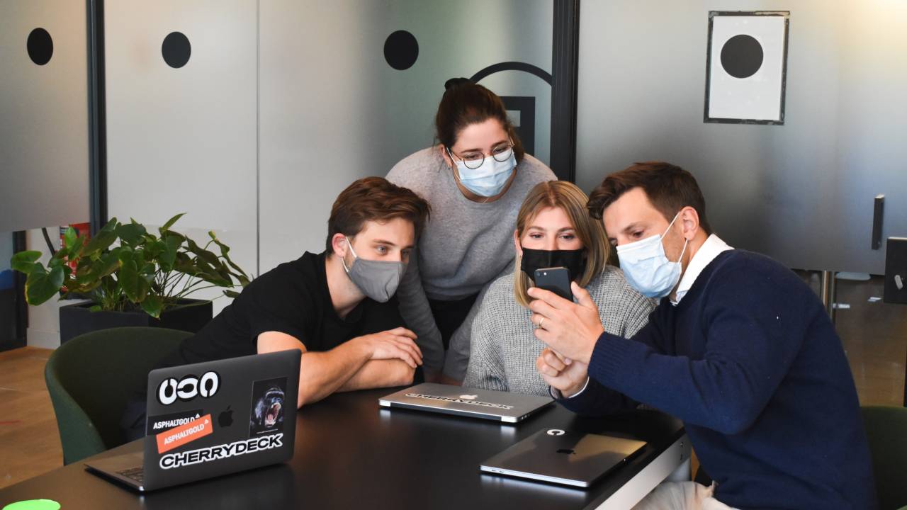 Imagem mostra dois homens brancos e duas mulheres brancas em uma mesa de reunião. Todos usam máscara de proteção contra a covid e olham para o celular de um dos homens.