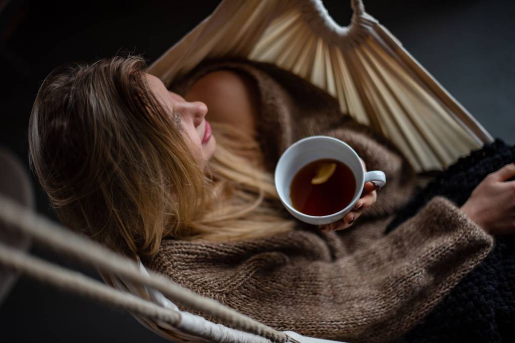 Imagem mostra uma mulher loira sentada em uma rede com uma xícara de chá na mão.
