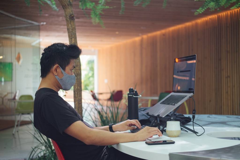 Imagem mostra um homem jovem, oriental, de máscara trabalhando em um notebook