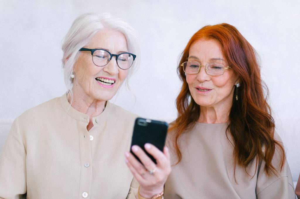 Imagem mostra duas mulheres brancas com mais de 60 anos. Uma delas tem cabelo grisalho e a outra é ruiva. Ambas estão olhando para um celular e sorrindo.