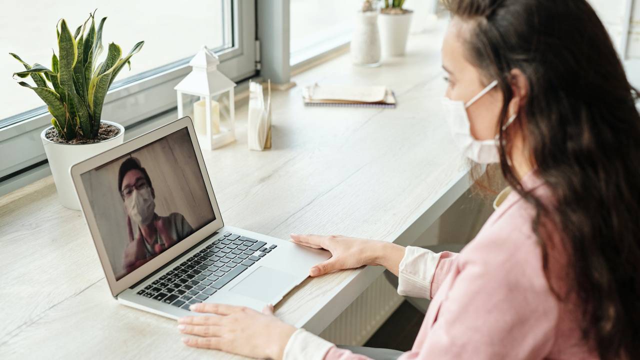 Imagem mostra uma mulher de máscara, de cabelos longos e escuros, conversando por meio de uma videochamada com um homem que também veste uma máscara.