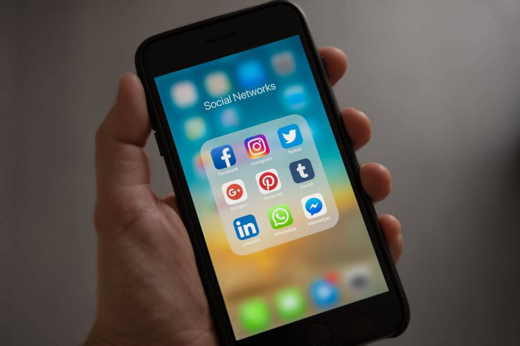 A imagem mostra uma mão segurando um smartphone, na tela estão dispostos ícones de redes sociais