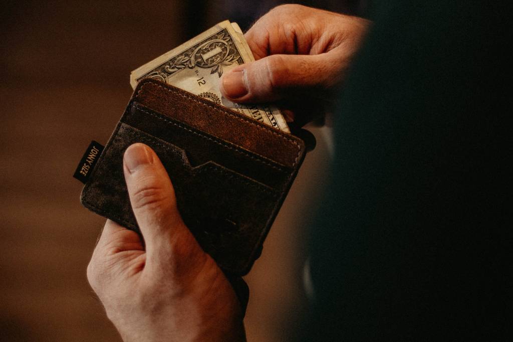 Imagem mostra duas mãos segurando uma carteira marrom cheia de dólares.