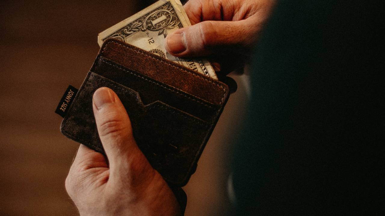 Imagem mostra duas mãos segurando uma carteira marrom cheia de dólares.