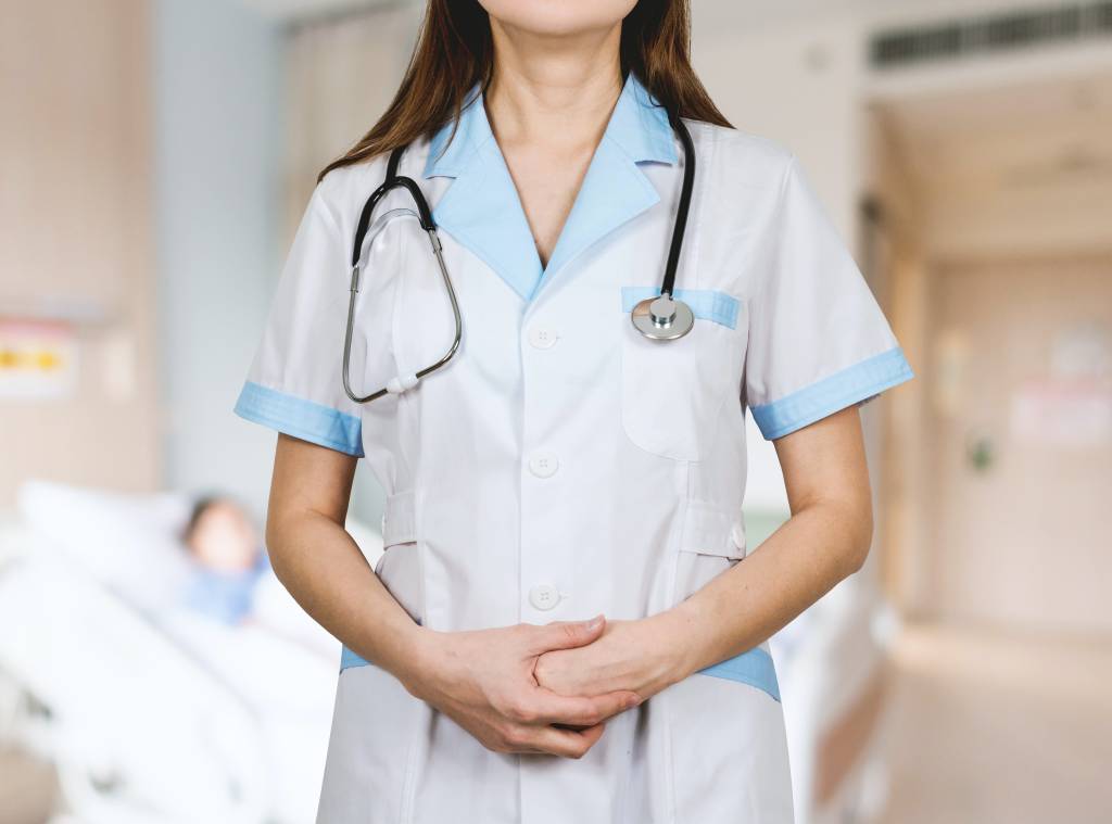 Imagem mostra uma mulher branca, de cabelos longos e escuros, com jaleco médico e estetoscópio no pescoço