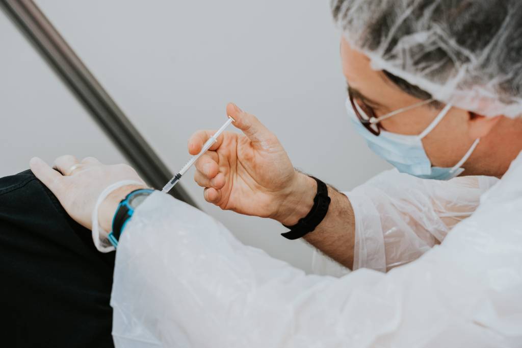 Imagem mostra uma enfermeira de óculos, touca e máscara aplicando uma vacina