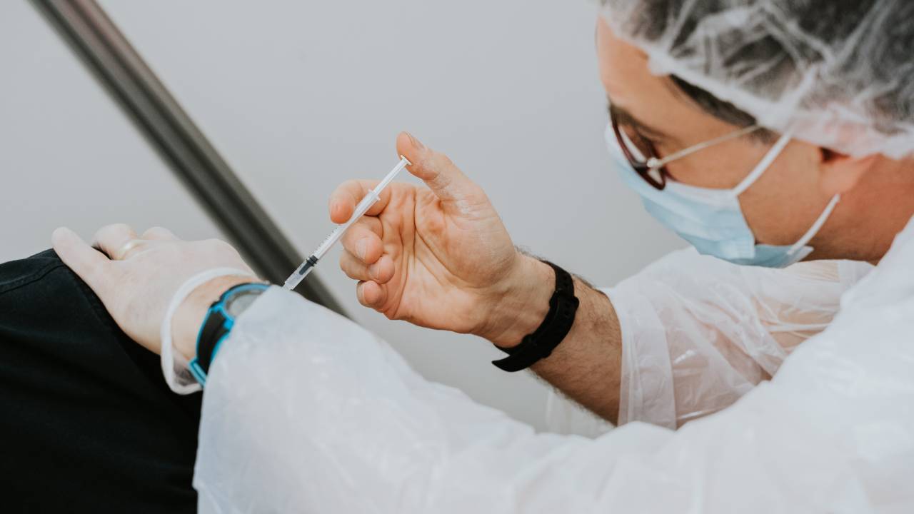Imagem mostra uma enfermeira de óculos, touca e máscara aplicando uma vacina