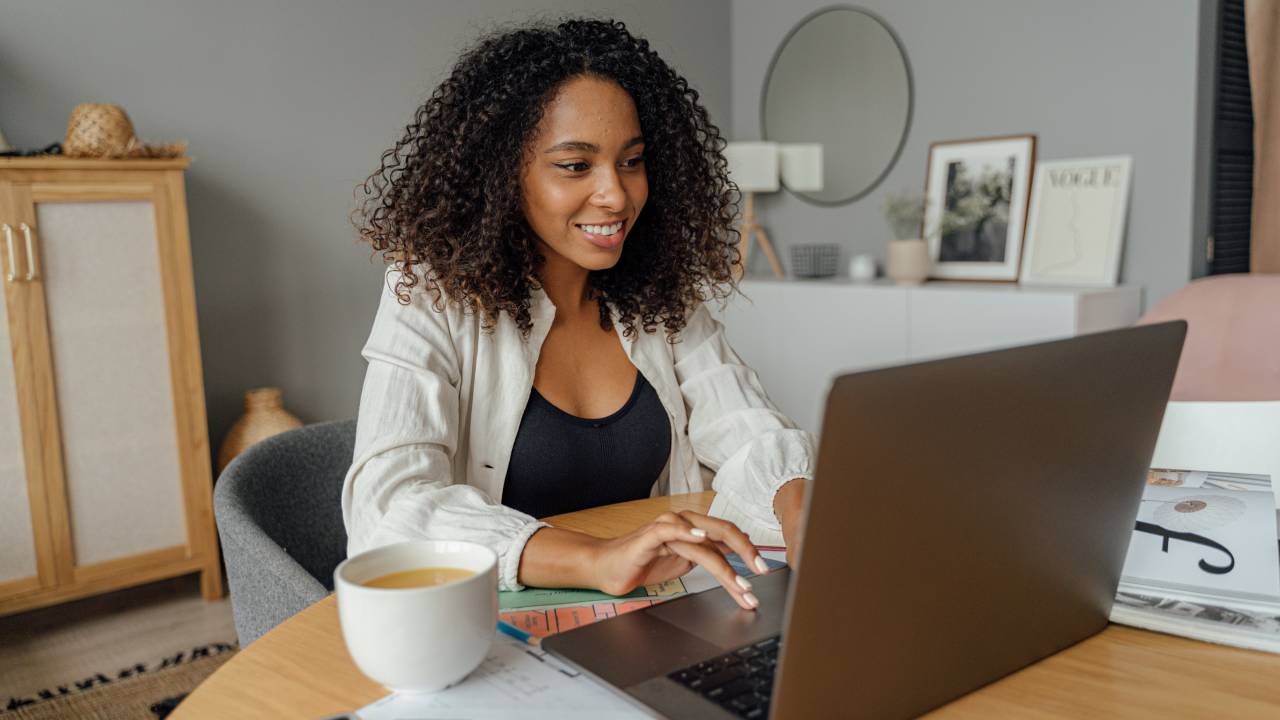 Imagem mostra uma mulher negra de cabelos afro sorrindo em frente a um notebook. Ela está trabalhando em casa.