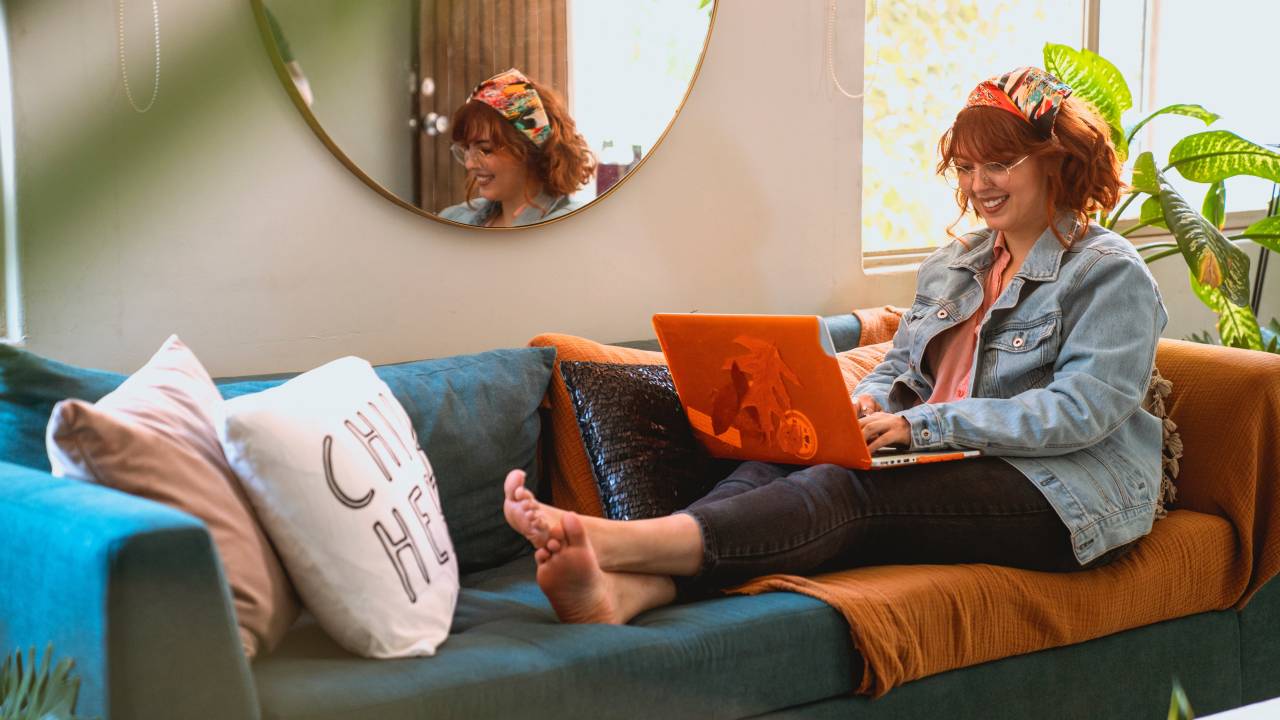 Imagem mostra mulher branca, de cabelos ruivos, sentada em um sofá azul usando o notebook