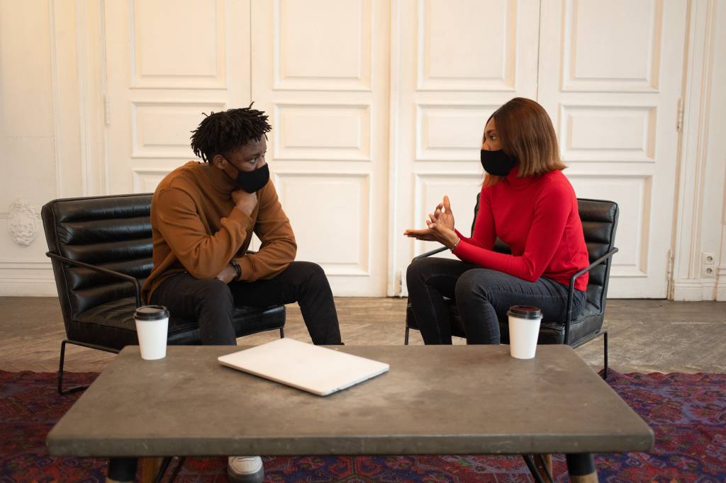 Imagem mostra um homem e uma mulher, ambos negros, conversando. Eles usam máscaras de proteção e cada um está sentado em uma poltrona.