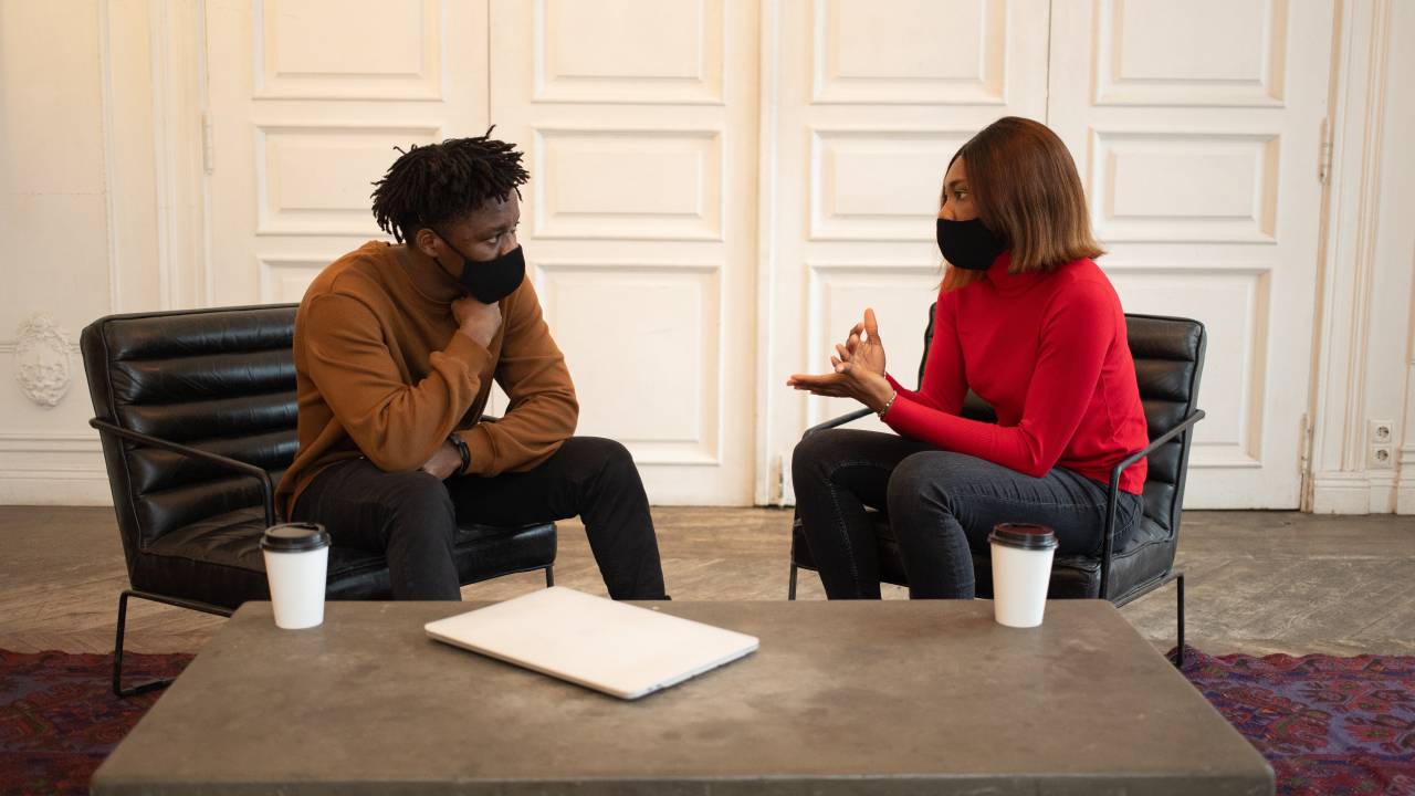 Imagem mostra um homem e uma mulher, ambos negros, conversando. Eles usam máscaras de proteção e cada um está sentado em uma poltrona.