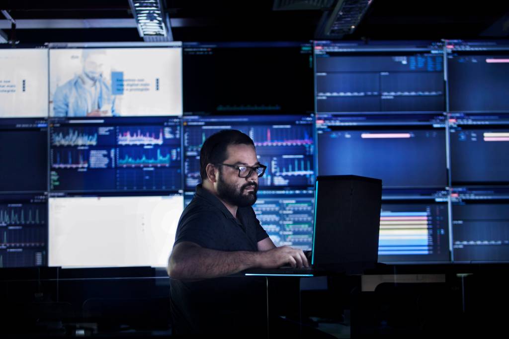 A imagem mostra Marcus Garcia mexendo no computador em um ambiente cheio de telas azuladas.