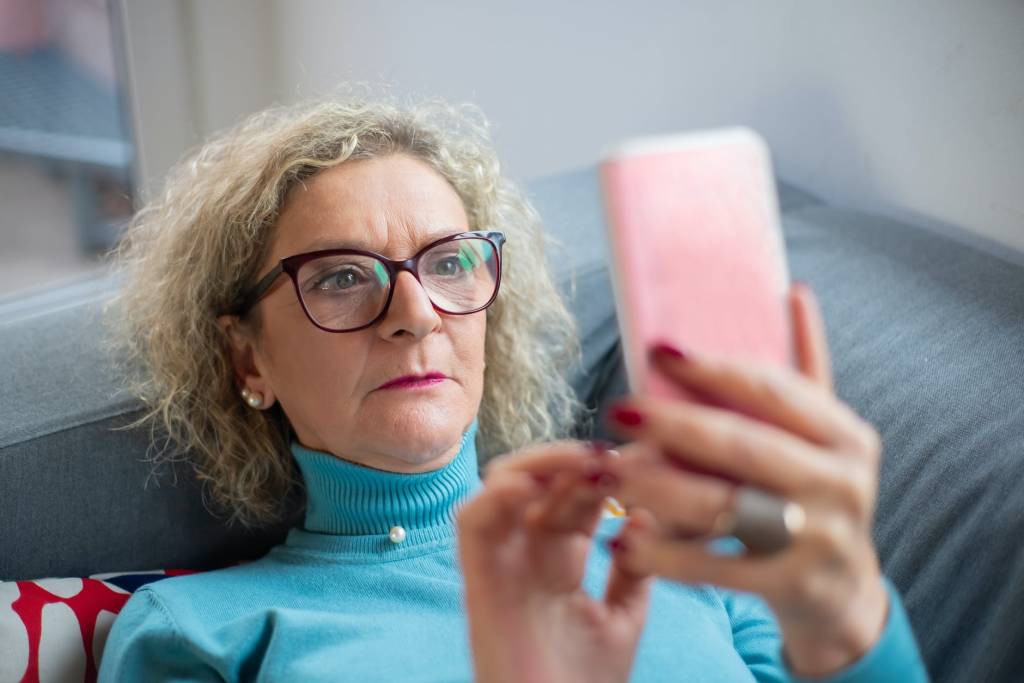 Imagem mostra uma mulher de cabelos grisalhos, com óculos, olhando para a tela de um smartphone