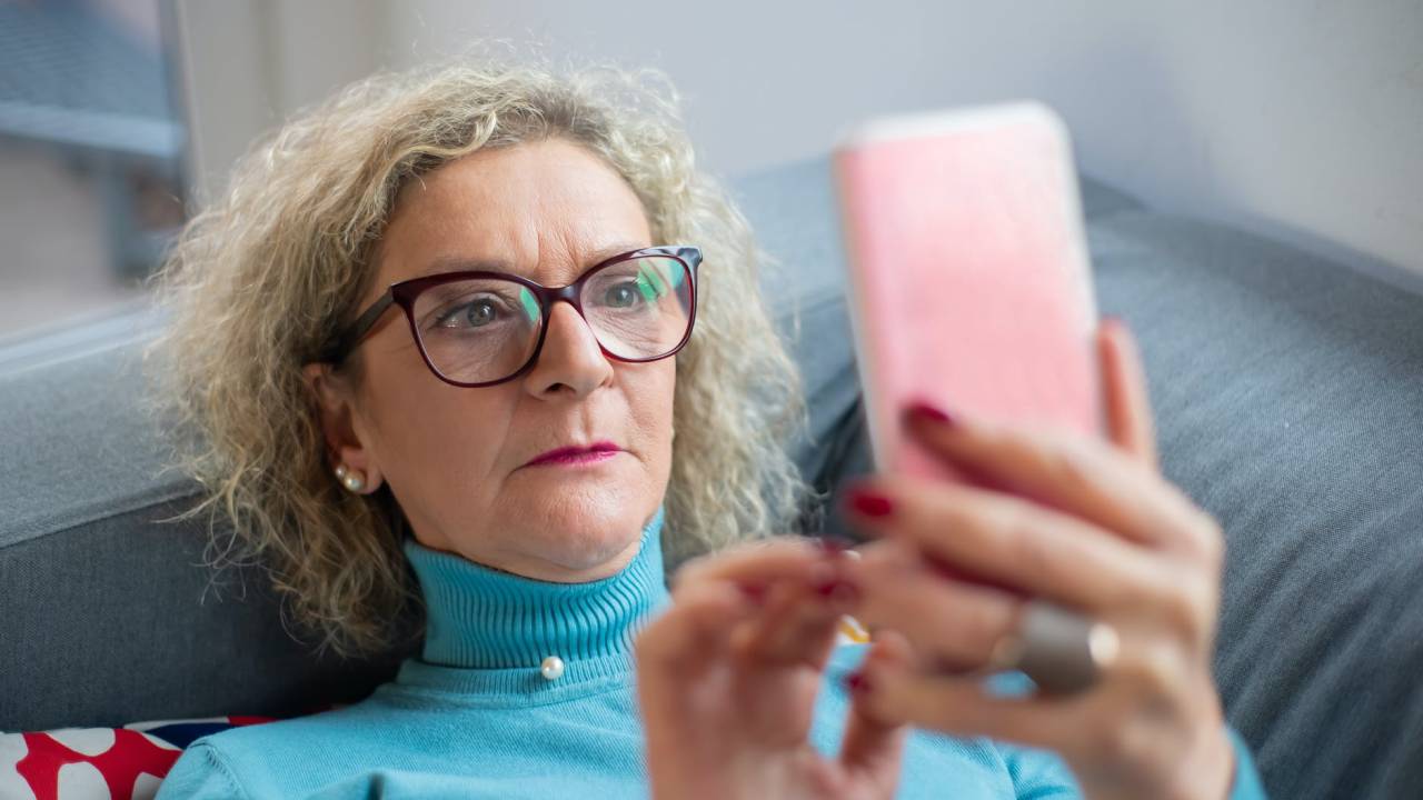 Imagem mostra uma mulher de cabelos grisalhos, com óculos, olhando para a tela de um smartphone