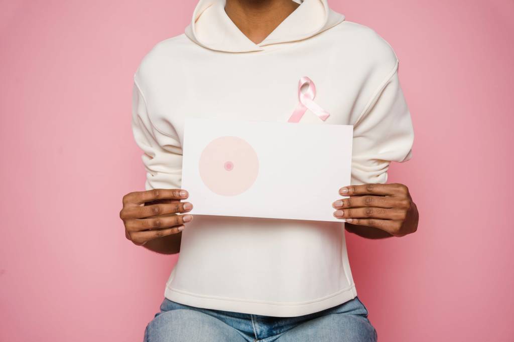 Imagem mostra uma mulher, sobre um fundo rosa, vestindo um moletom e segurando a imagem de uma mama. No moletom, está uma fita cor de rosa, que simboliza o Outubro Rosa.
