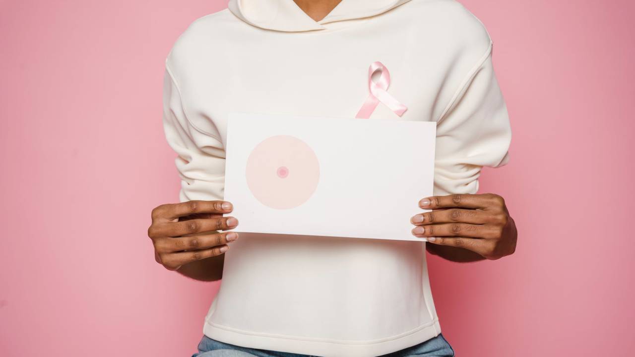 Imagem mostra uma mulher, sobre um fundo rosa, vestindo um moletom e segurando a imagem de uma mama. No moletom, está uma fita cor de rosa, que simboliza o Outubro Rosa.