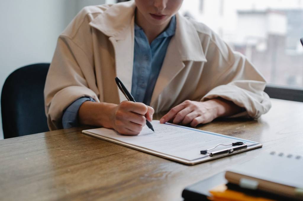 Imagem mostra uma mulher sentada em frente a uma mesa preenchendo uma ficha à caneta.