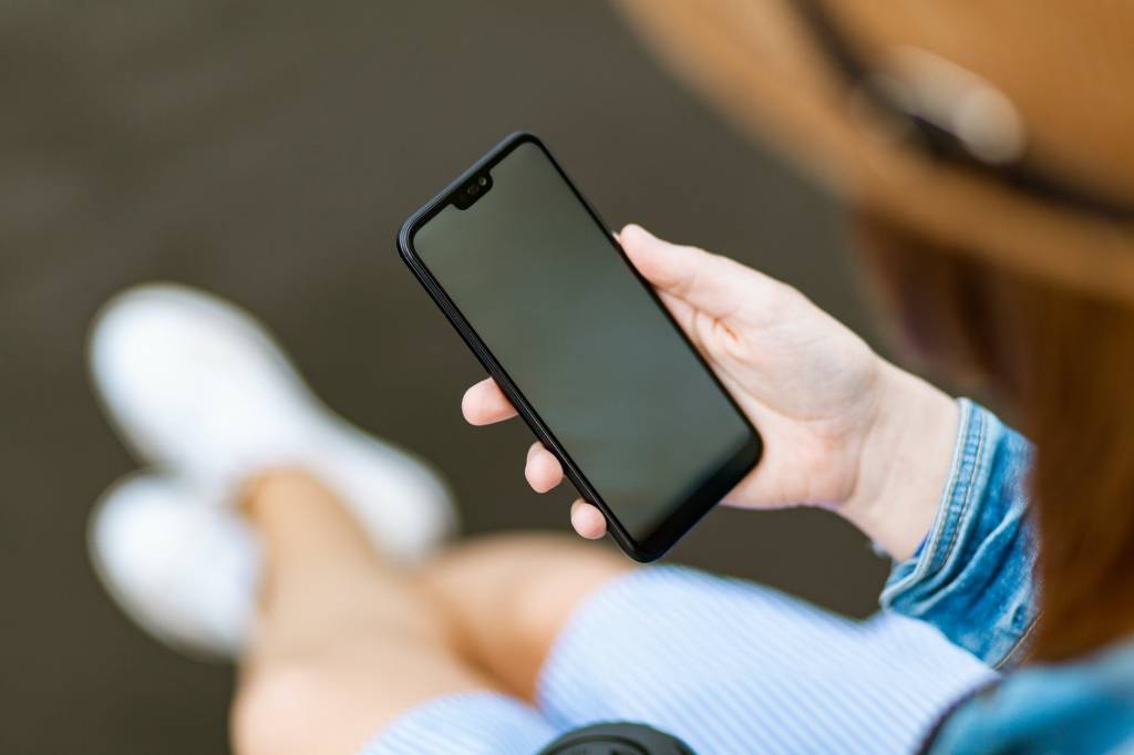 Imagem mostra uma pessoa sentada com um celular na mão. O foco é no smartphone, sem mostrar o rosto de quem o segura.