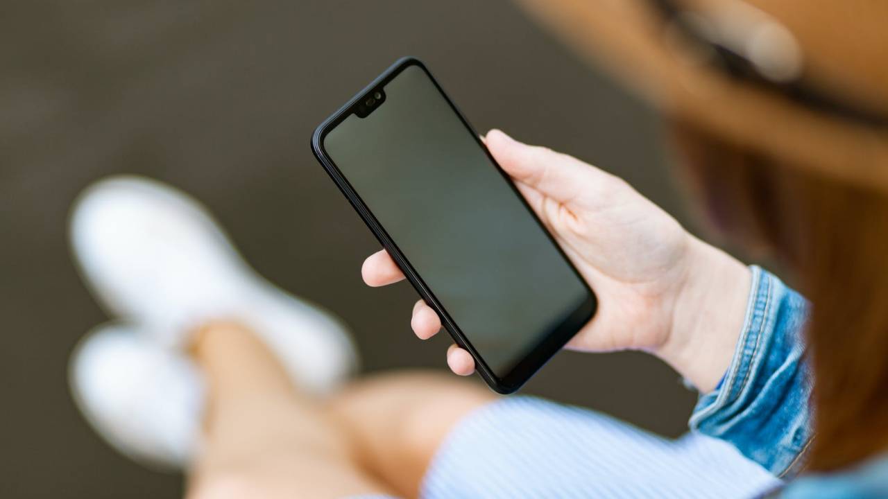 Imagem mostra uma pessoa sentada com um celular na mão. O foco é no smartphone, sem mostrar o rosto de quem o segura.
