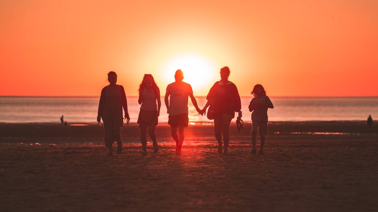 Imagem mostra as silhuetas de cinco pessoas andando em uma praia no pôr-do-sol
