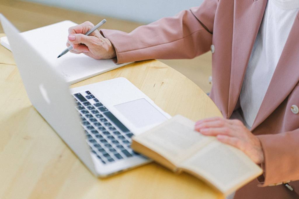 Imagem mostra as mãos de uma mulher mais velha, vestindo um terno rosa, fazendo anotações em um caderno enquanto mexe no computador e lê um livro