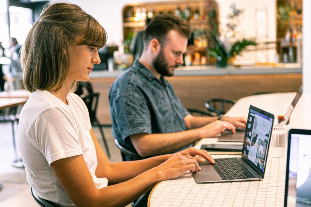Imagem mostra uma mulher e um homem trabalhando um ao lado do outro no computador. Ela aparece em primeiro plano, ele em segundo.