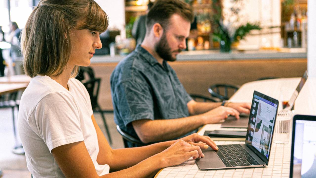 Imagem mostra uma mulher e um homem trabalhando um ao lado do outro no computador. Ela aparece em primeiro plano, ele em segundo.