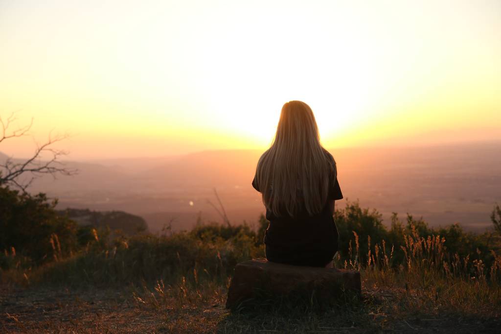 Imagem mostra uma mulher loira, de costas, sentada em um banco olhando um horizonte no pôr-do-sol