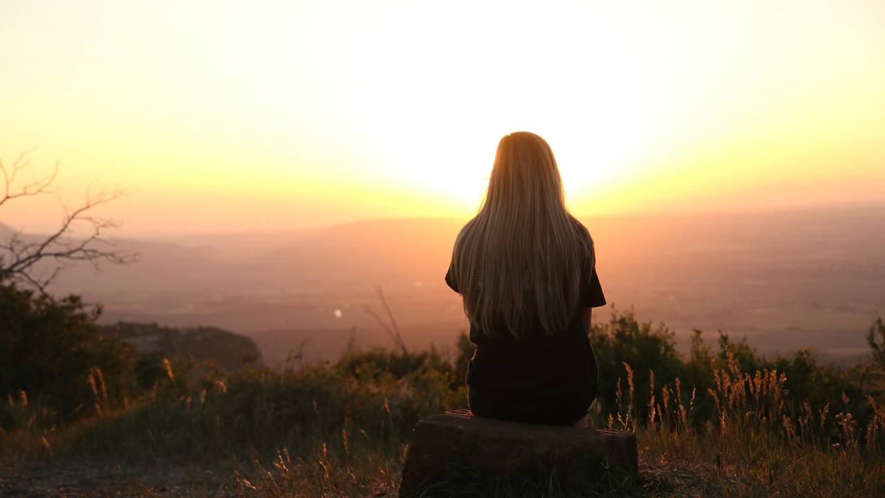 Imagem mostra uma mulher loira, de costas, sentada em um banco olhando um horizonte no pôr-do-sol