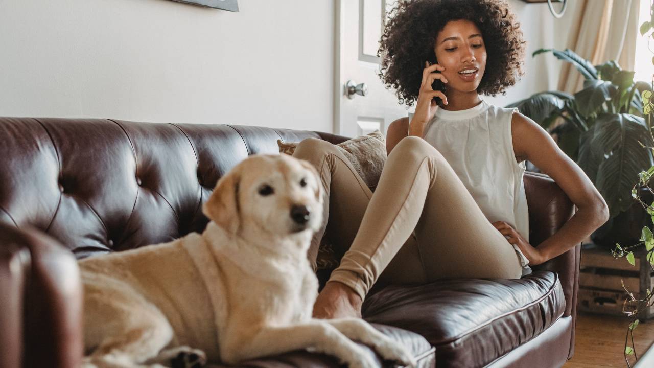 Imagem mostra uma mulher negra, de blusa branca e cabelos escuros cacheados. Ela está em um sofá, ao lado de um cachorro bege, falando no celular.