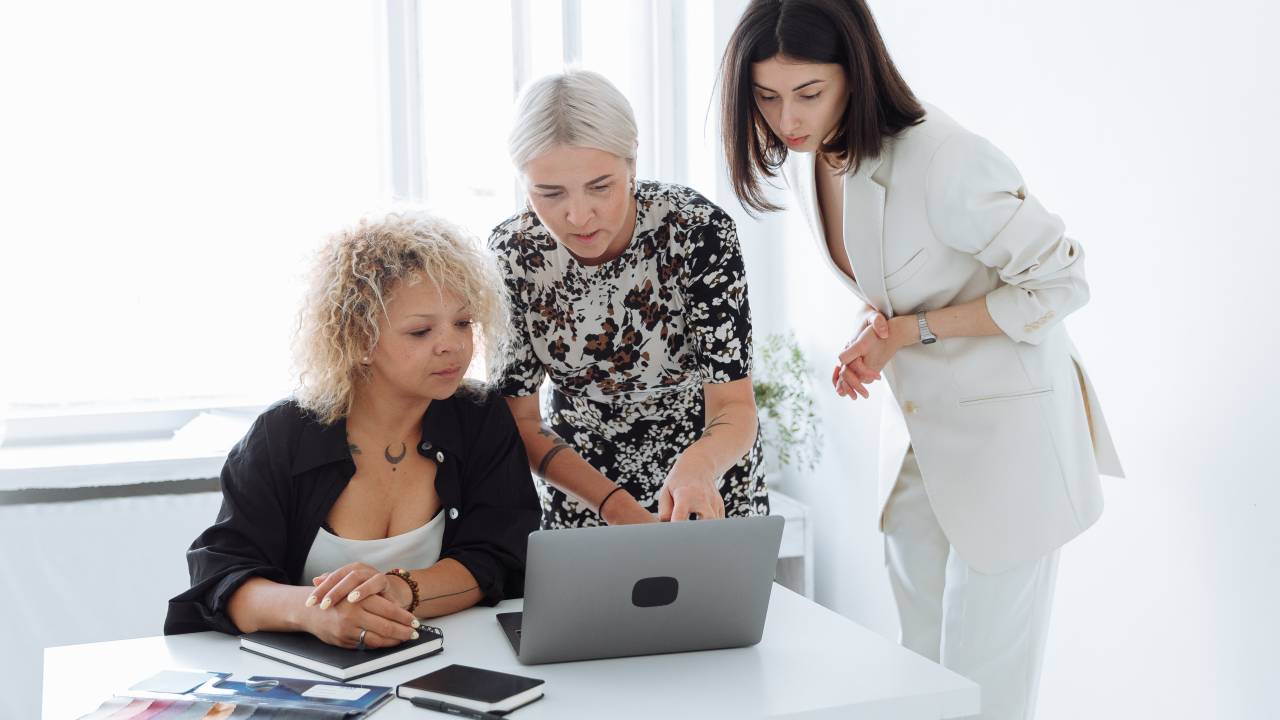 Imagem mostra três mulheres em uma mesa de reunião, em volta do computador. Uma delas é jovem, branca e tem cabelos lisos e escuros, a outra é mais velha, branca e tem cabelos grisalhos e a terceira é negra e tem cabelos crespos loiros.