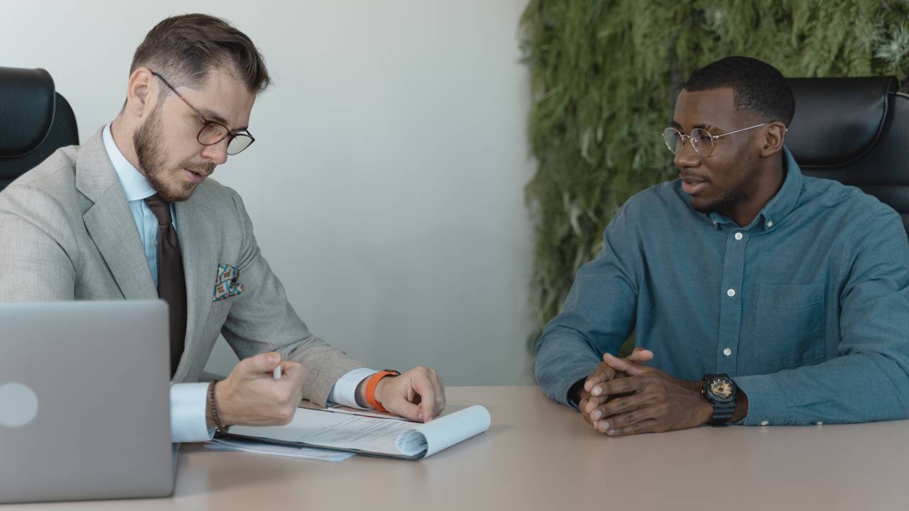 Imagem mostra dois homens em uma entrevista de emprego. O entrevistador é branco, usa óculos e blazer cinza e lê algo em um papel. O entrevistado é negro e usa uma camisa azul.