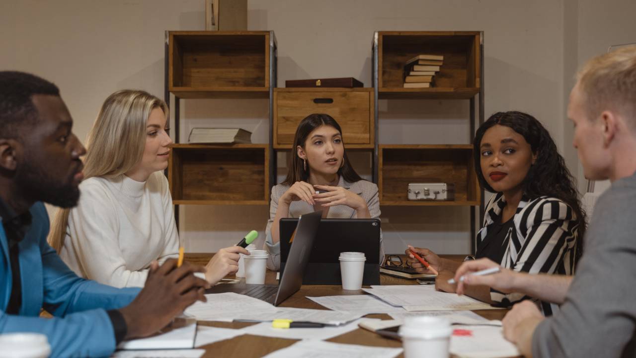 Imagem mostra cinco pessoas conversando em uma mesa de reunião. São duas mulheres brancas, uma mulher negra, um homem negro e um homem branco.
