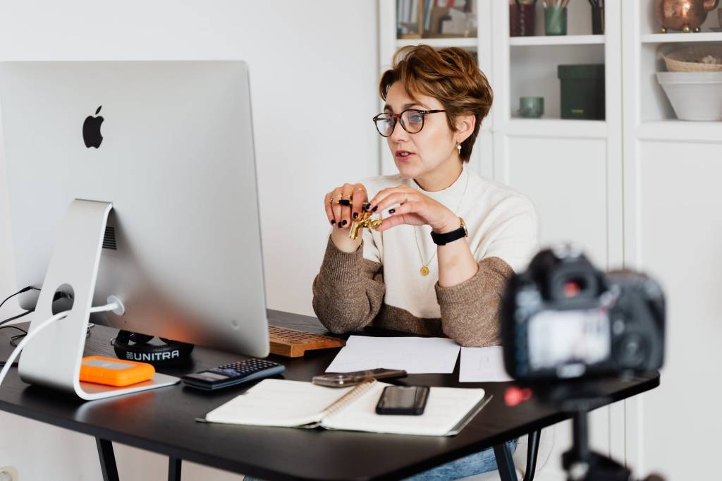 Imagem mostra uma mulher interagindo com alguém através do computador. Ela está sentada em uma mesa de trabalho e, em primeiro plano, aparece uma câmera aparentemente gravando a situação