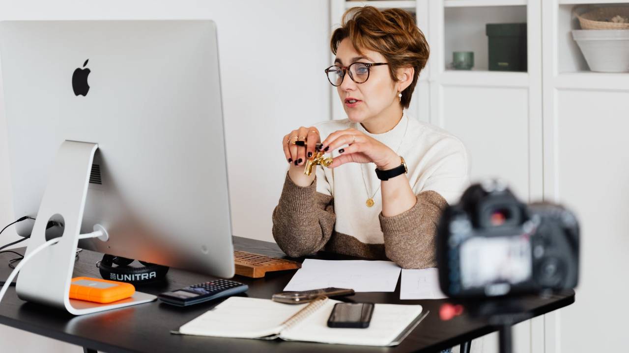 Imagem mostra uma mulher interagindo com alguém através do computador. Ela está sentada em uma mesa de trabalho e, em primeiro plano, aparece uma câmera aparentemente gravando a situação