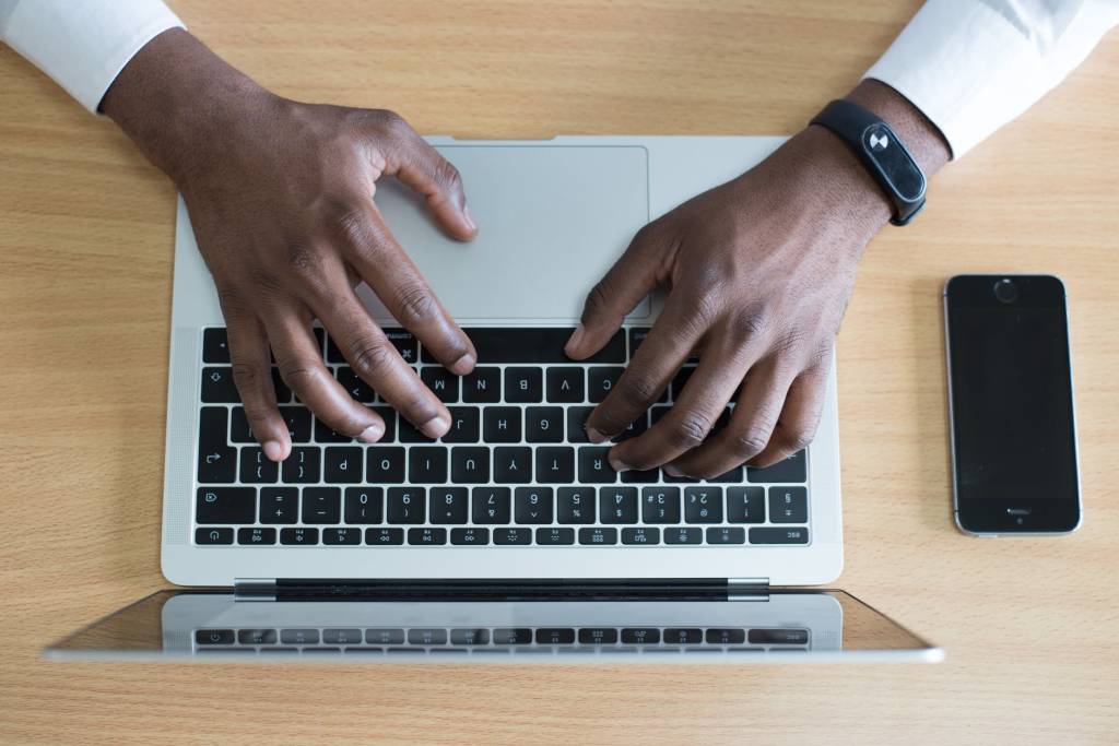Imagem mostra um teclado de notebook e duas mãos sobre ele, digitando. No pulso de uma delas, há um smartwatch