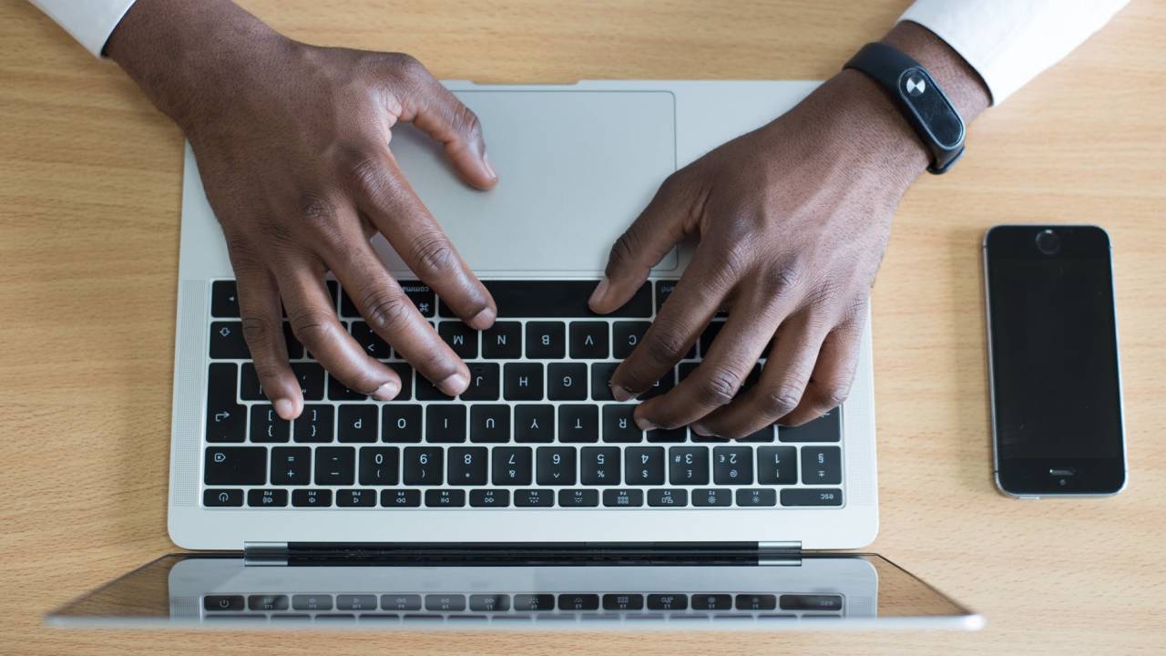 Imagem mostra um teclado de notebook e duas mãos sobre ele, digitando. No pulso de uma delas, há um smartwatch